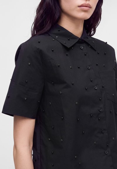 Femme portant une chemise noire à manches courtes avec col et boutons, décorée de petites perles noires réparties uniformément sur le tissu.