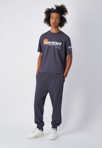 Camiseta de algodón gris oscuro con gráfico de "PRESENTE" en blanco y naranja, combinada con pantalones de chándal a juego y zapatillas blancas. Textura suave, corte relajado.