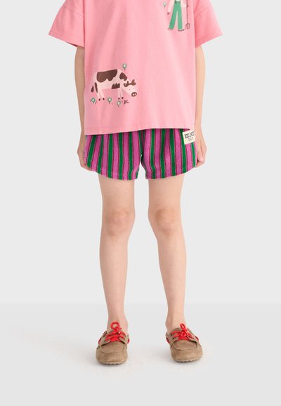 Enfant portant un t-shirt rose avec un motif d'animaux de la ferme, un short rayé violet et vert, et des chaussures marron avec des lacets rouges, debout sur un fond blanc.