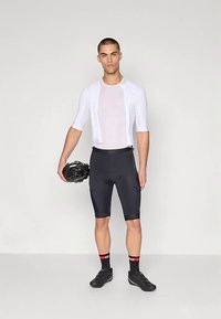 Νεαρός άνδρας που φοράει λευκό ποδηλατικό μπλουζάκι, μαύρες ποδηλατικές βερμούδες, μαύρα ποδηλατικά παπούτσια και κρατάει ένα μαύρο κράνος σε απλό φόντο.