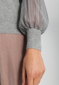 Szary sweter dzianinowy z przezroczystymi puff sleeves, prążkowanym mankietem i teksturowanym wzorem. Miękka tkanina kontrastuje z gładką, półprzezroczystą siateczką.