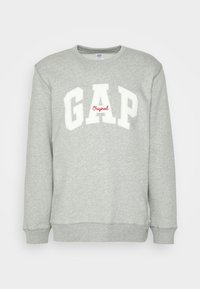 Sudadera gris claro con letras blancas de GAP y pequeño texto rojo "Original" centrado en el pecho, con mangas largas y puños acanalados.