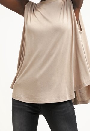 Beżowy top bez rękawów z luźnym, zwiewnym materiałem, noszony przez osobę unoszącą ramiona, w połączeniu z dopasowanymi czarnymi dżinsami.