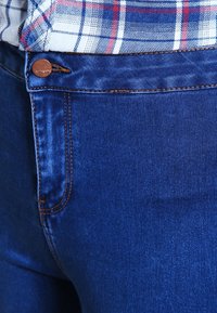 Tmavě modré džínové kalhoty s hladkou texturou, vybavené hnědým kovovým knoflíkem a oranžovým šitím podél kapes a pasu.