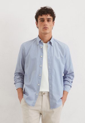 Button-down shirt with blue and white stripes, long sleeves, soft cotton fabric, paired with a white crew-neck t-shirt and beige trousers.