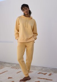 Gelbes Hoodie mit Kordeln, kombiniert mit passenden Sweatpants. Glatter Stoff, lockerer Schnitt und gerippte Bündchen. Minimalistisches Design.