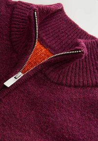 Paarse gebreide trui met een hoge kraag, voorzien van een halve rits die een oranje voering onthult. De stof lijkt zacht en textuur te hebben met ribbelaccenten.