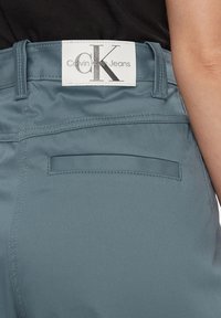 Pantaloni di tessuto azzurro chiaro con una consistenza liscia, dotati di un'etichetta bianca con "Calvin Klein Jeans" e una tasca posteriore.
