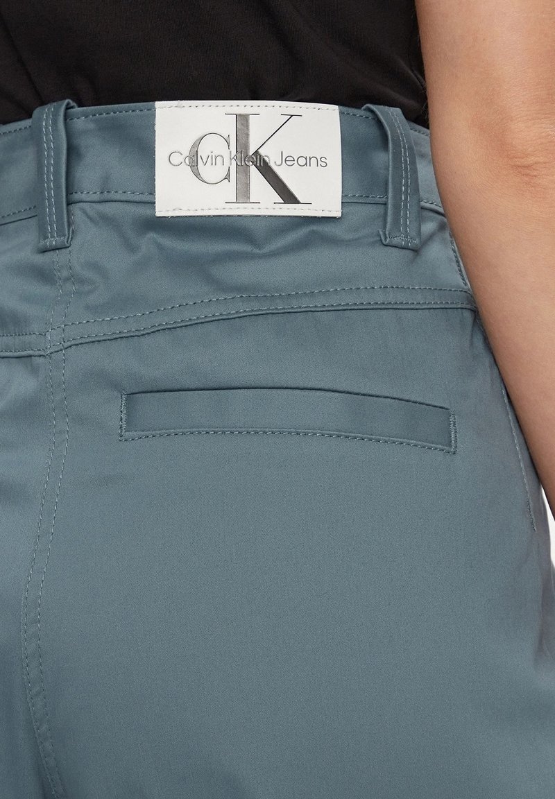 Pantaloni di tessuto azzurro chiaro con una consistenza liscia, dotati di un'etichetta bianca con "Calvin Klein Jeans" e una tasca posteriore.