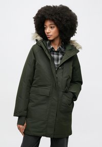 Ciemnozielona kurtka parka z kapturem wyściełanym futrem, zapinana na zamek, z dwoma przednimi kieszeniami. Nosi się ją na czarno-białej koszuli w kratę o luźnym kroju.