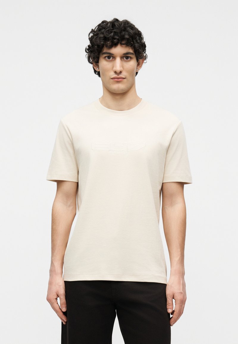 Νεαρός άνδρας με σγουρά μαλλιά, φορώντας απλή μπεζ κοντομάνικη μπλούζα και μαύρο παντελόνι, στέκεται μπροστά σε λευκό φόντο.