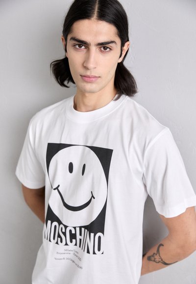 Hvit bomull T-skjorte med et stort svart smiley-fjes grafikk og "MOSCHINO" logo i fet skrift nedenfor. Korte ermer og rund hals.