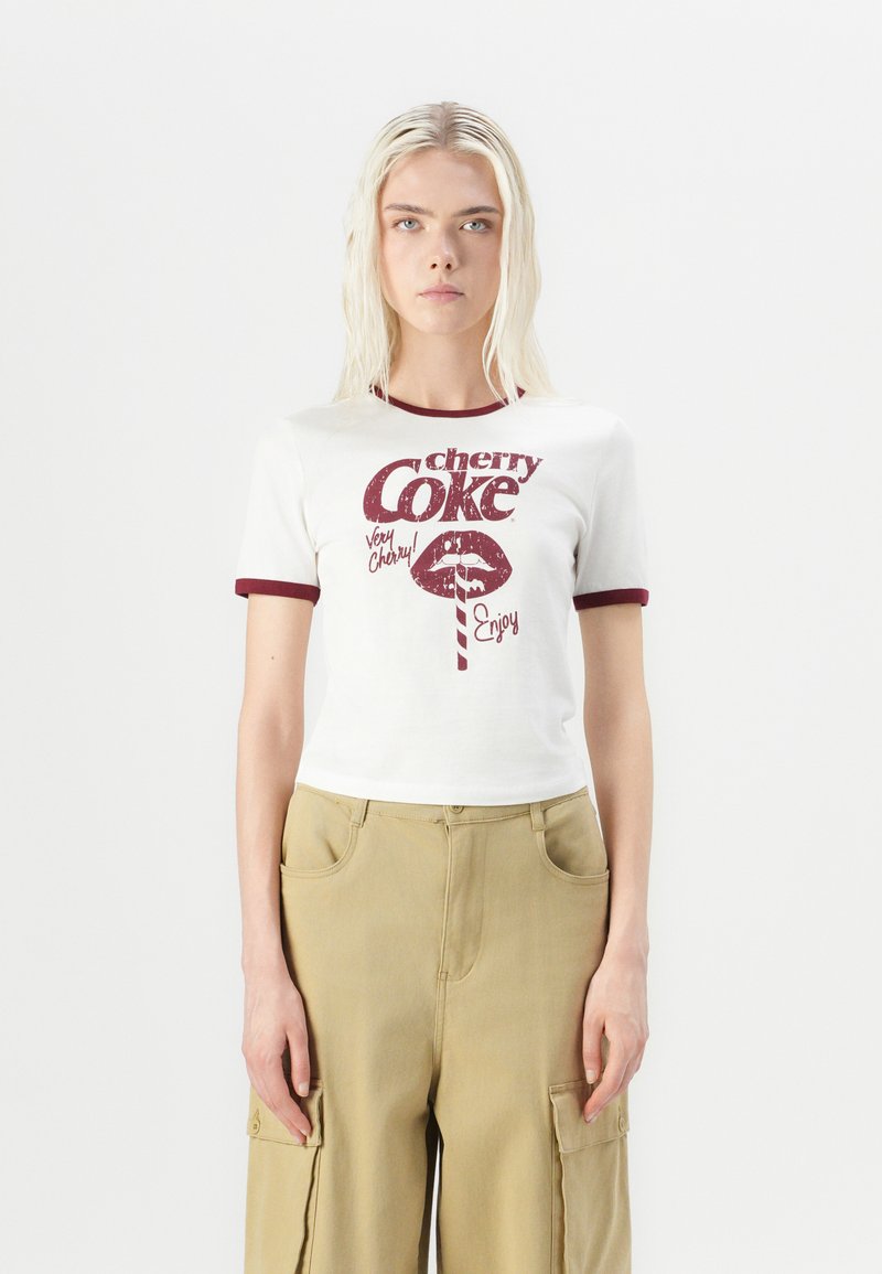 White cropped t-shirt with maroon trim, featuring a vintage-style "cherry Coke" graphic and text. Model wears khaki cargo pants.