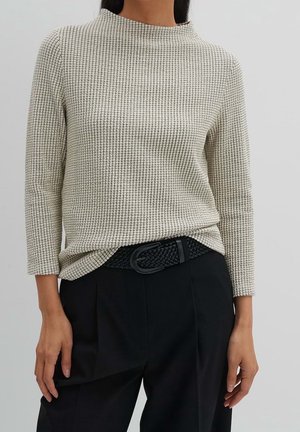 Mujer con camiseta beige texturizada de manga larga metida dentro de pantalones negros con un cinturón negro trenzado, de pie frente a un fondo claro y liso.