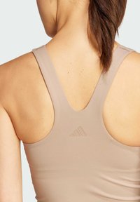Camiseta deportiva de color beige claro con un escote profundo en la espalda, tirantes finos y logotipo de la marca en relieve. Textura de tela suave, diseño minimalista.