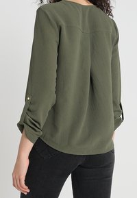 Blusa leggera verde oliva a maniche lunghe con polsini risvoltati, tessuto strutturato e orlo arrotondato. Include dettagli di cucitura sottili.