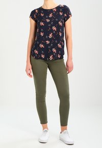 Námořnická květinová blůza s růžovými akcenty, krátkými rukávy a kulatým výstřihem, kombinovaná s olivově zelenými úzkými džíny a bílými teniskami.