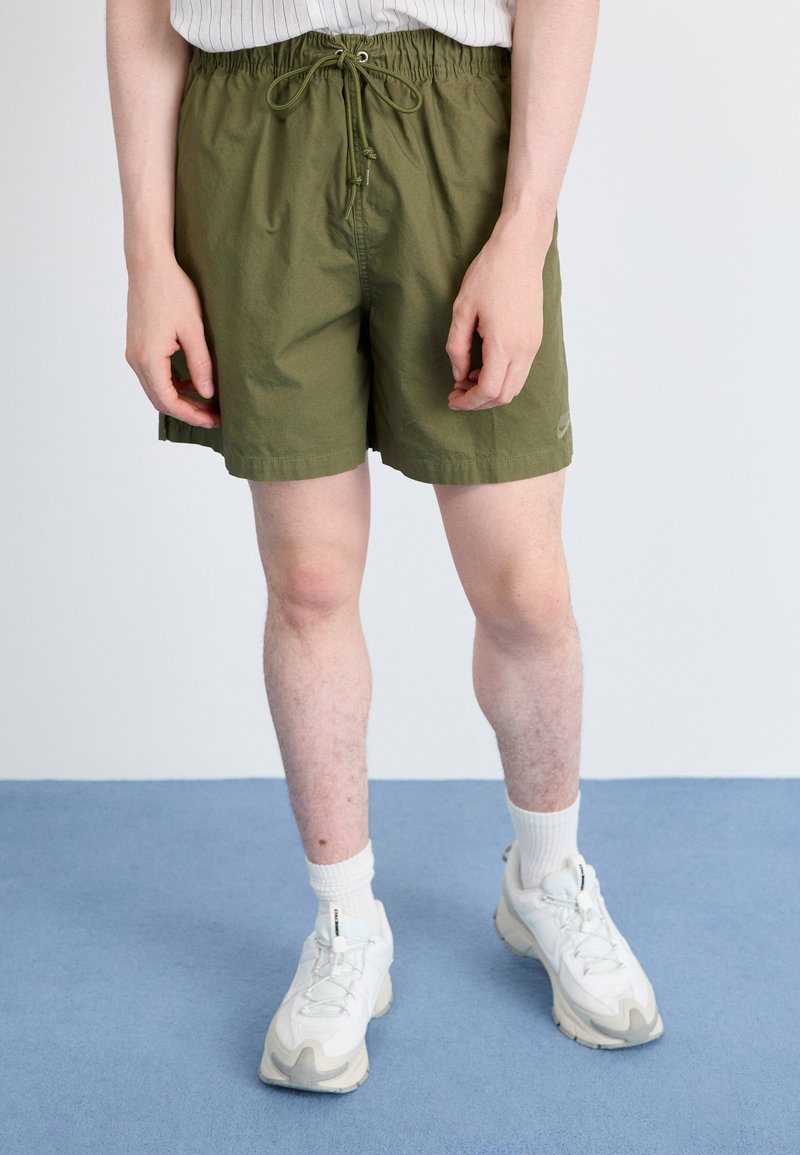 Calções de banho verde-azeitona com cintura elástica e cordão. Apresentam bolsos laterais e um pequeno logótipo na bainha. Usados com ténis brancos e meias.