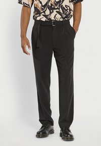 Pantalons noirs ajustés avec une finition lisse, dotés d'une ceinture à nouer sur le côté, d'un devant plissé et d'une coupe décontractée, associés à des chaussures noires à lacets.