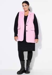 Rosa ärmellose Blazer mit zwei Fronttaschen und dunklen Knöpfen, über ein schwarzes, glitzerndes Kleid getragen und kombiniert mit kniehohen schwarzen Stiefeln.
