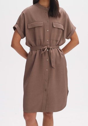 Mujer con vestido camisa marrón de manga corta hasta la rodilla, con botones delanteros, bolsillos en el pecho y cinturón atado a la cintura, de pie con las manos en las caderas.