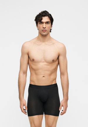 Młody mężczyzna o ciemnych kręconych włosach, ubrany w czarne bokserki, stojący na jednolitym jasnoszarym tle, zwrócony przodem.