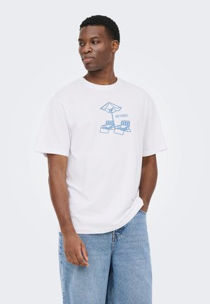 Homme portant un t-shirt blanc avec une impression bleue de deux chaises longues sous un parasol et le mot «RETRAITÉ», associé à un jean bleu clair.