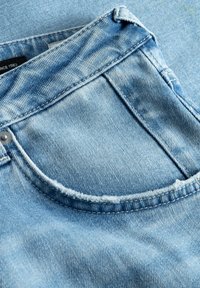 Ljusblå denimjeans med en böjd ficka med slitna kanter och en silverknapp, visar en mjuk textur och klassiska sömnadsdetaljer.