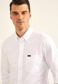 Camisa blanca de botones hecha de algodón, con cuello puntiagudo, un bolsillo en el pecho y un pequeño logo en navy en el bolsillo. Textura suave.