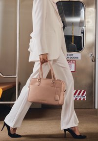 Ljust rosa läderhandväska med dubbla handtag, texturerad yta och detaljer i guld, bärs av en person i vit kostym och klackar.