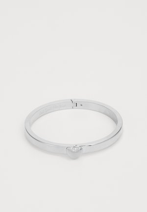 Bracelet jonc en métal argenté avec un petit fermoir en forme de cœur sur un fond clair uni.
