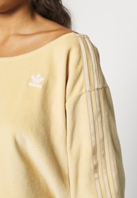 Gul sweatshirt i mjukt material, med en bred halsringning, tre vita vertikala ränder på ärmarna och en broderad logotyp på bröstet.