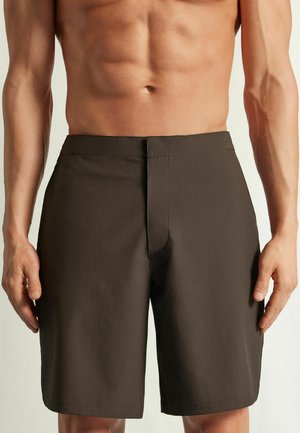 Männlicher Torso trägt einfarbige dunkelbraune, knielange Shorts mit flacher Vorderseite und ohne sichtbare Taschen, steht mit entspannten Armen an den Seiten.