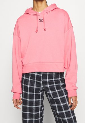 Persoon draagt een felroze Adidas hoodie met trekkoorden en zwart-witte geruite broek, staand tegen een effen achtergrond.