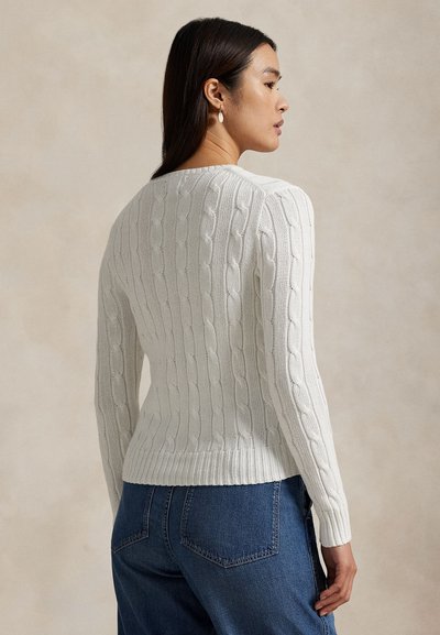 Vrouw van opzij, draagt een witte kabelgebreide trui en blauwe spijkerbroek tegen een effen beige achtergrond.