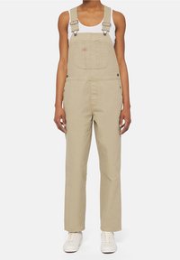 Salopette en coton beige avec des bretelles réglables, une poche avant et des accessoires en métal, associée à un débardeur blanc ajusté et des baskets blanches.