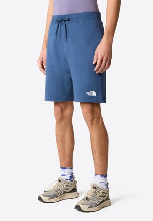 Shorts atletici in cotone blu con elastico in vita e cordoncino. Presentano un logo bianco sulla gamba sinistra. Indossati con scarpe da ginnastica beige e grigie.