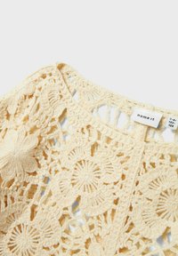 Top all'uncinetto di un leggero colore beige con motivi floreali e geometrici. Collo rotondo con dettagli a punto aperto. Etichetta visibile di lato.