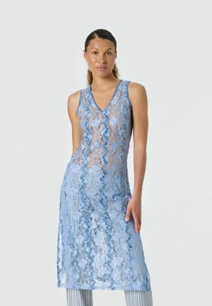 Femme portant une robe sans manches en dentelle florale bleu clair sur un pantalon rayé, debout devant un fond clair uni.