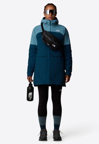 Vandafvisende blå og teal jakke, sort talje taske, sorte leggings, blå sokker, grå vandrestøvler, holder en sort vandflaske.