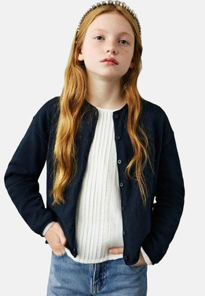 Tmavě modrý kardigan z měkké látky, s kulatým výstřihem, dlouhými rukávy a zapínáním na knoflíky, vrstvený přes bílý žebrovaný svetr.