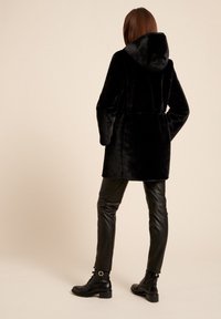 Abrigo de piel sintética negro con capucha, cintura ceñida y mangas largas, combinado con pantalones de cuero negros y botas de tobillo. Tela texturada visible.