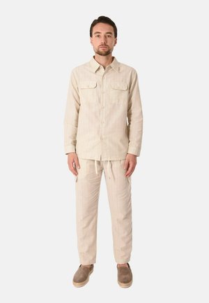 Uomo in piedi di fronte, indossa una camicia beige con bottoni e tasche sul petto, pantaloni abbinati e scarpe marroni slip-on.