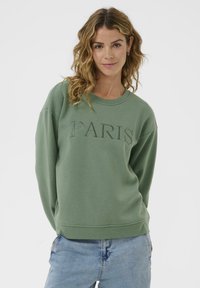 Sweatshirt vert avec "PARIS" brodé dans un fil assorti. Présente un col rond, des épaules tombantes et des poignets côtelés. Texture lisse.