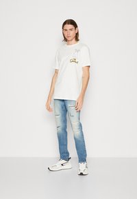 Camiseta blanca de manga corta con un gráfico de dibujos animados y texto en el lado izquierdo del pecho, combinada con jeans desgastados de color azul claro y zapatillas blancas.