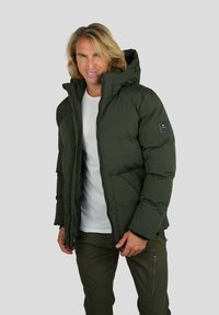 Homme aux cheveux blonds portant une veste matelassée vert foncé et un pantalon cargo assorti, se tenant devant un fond gris clair.