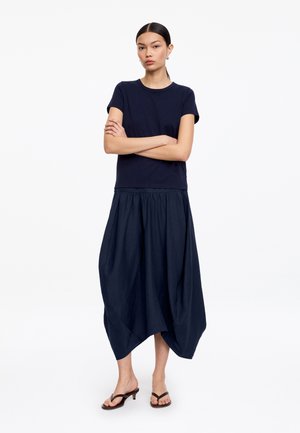 Mujer de pie con los brazos cruzados, vistiendo una camisa de manga corta azul marino, pantalones anchos azules marinos recortados y sandalias marrones con tacón.