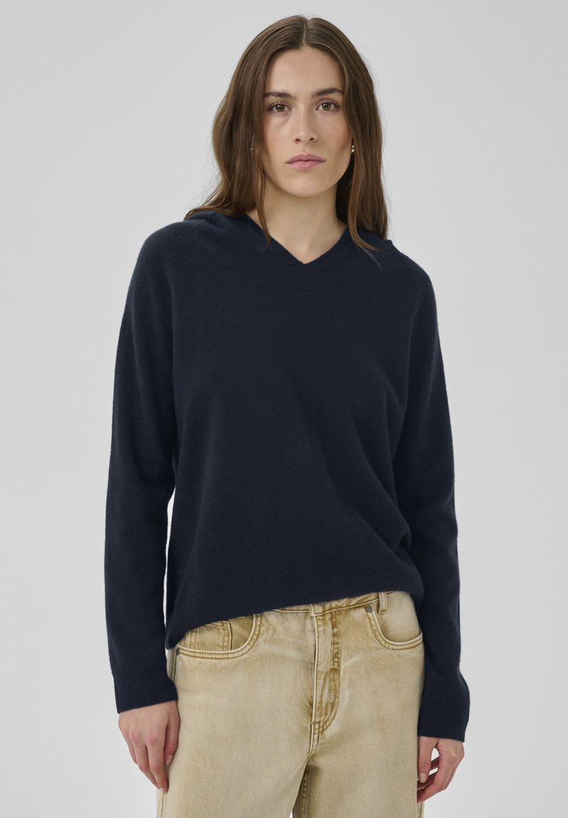 Marineblauer V-Ausschnitt-Pullover aus weichem Stoff mit langen Ärmeln und gerippten Bündchen, kombiniert mit hellbeigen hochgeschnittenen Hosen.