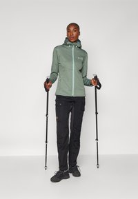 Zaļš kapuci ar rakstainu dizainu un gaišu rāvējslēdzēju, apvienots ar melnām cargo biksēm un pārgājienu nūjām. Iedarbīgas kurpes noslēdz āra apģērba komplektu.