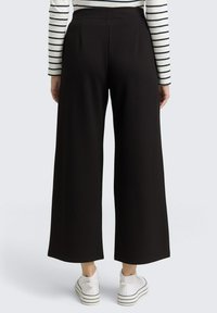 Pantalons larges noirs en tissu texturé, dotés d'une taille haute et de plis, associés à des baskets blanches avec des rayures noires.